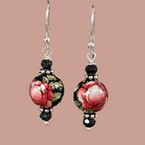 a black round vintage bead is painted with a red rose and green leaves and hanging from sterling silver ear wires and bordered by small black roundel beads and silver spacers